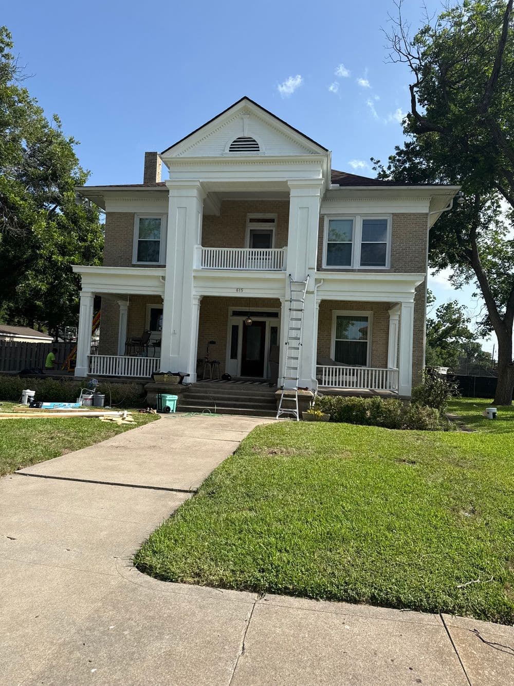 Historic two-story brick house with porch and ladder, surrounded by greenery and clear blue sky.