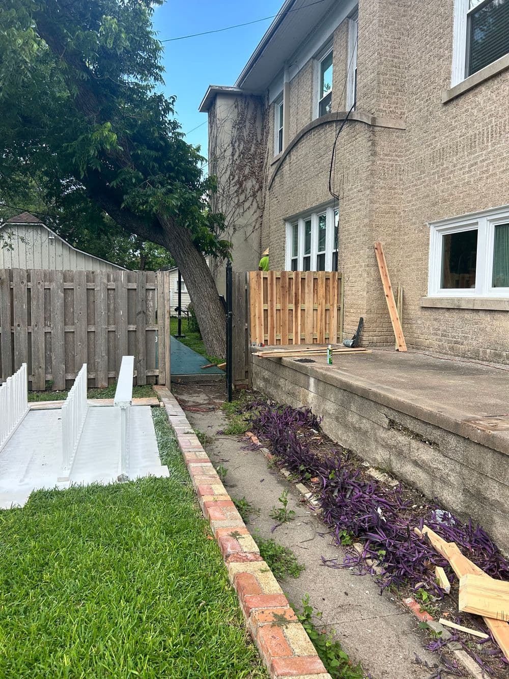 Renovation scene showing a house exterior, wooden fence, and garden with purple plants.