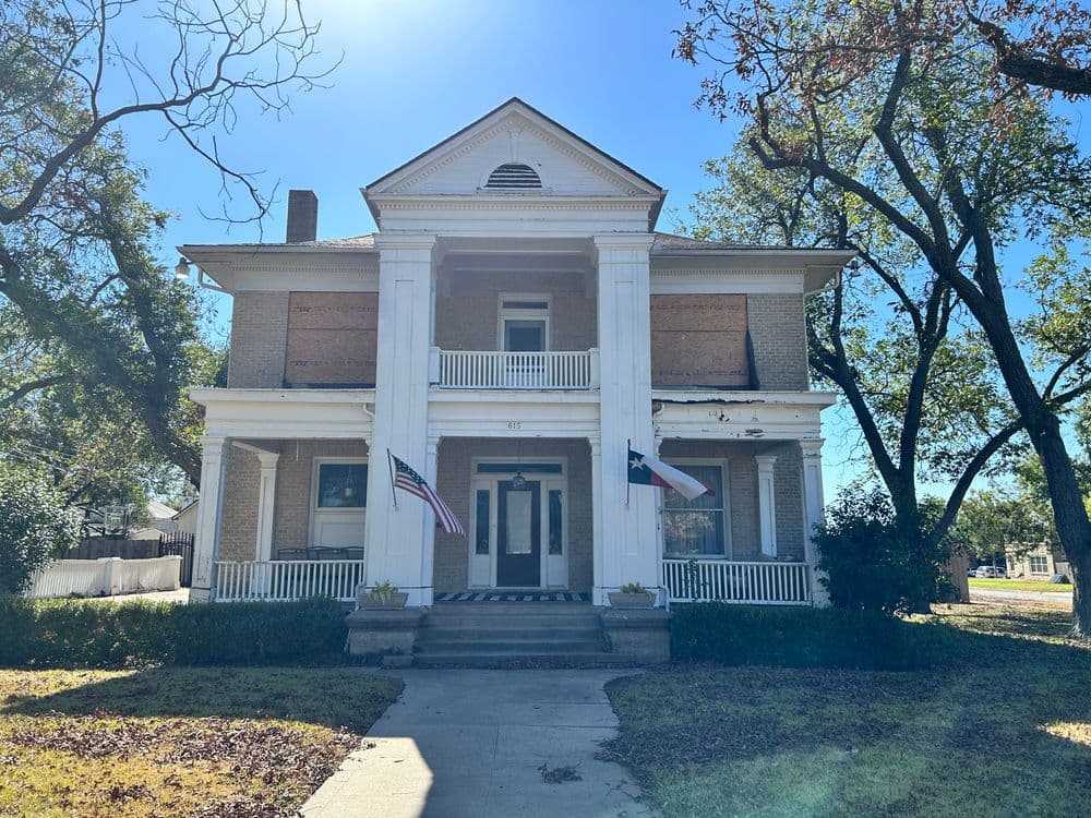 Historic two-story home with columns, American flags, and boarded windows under blue sky.