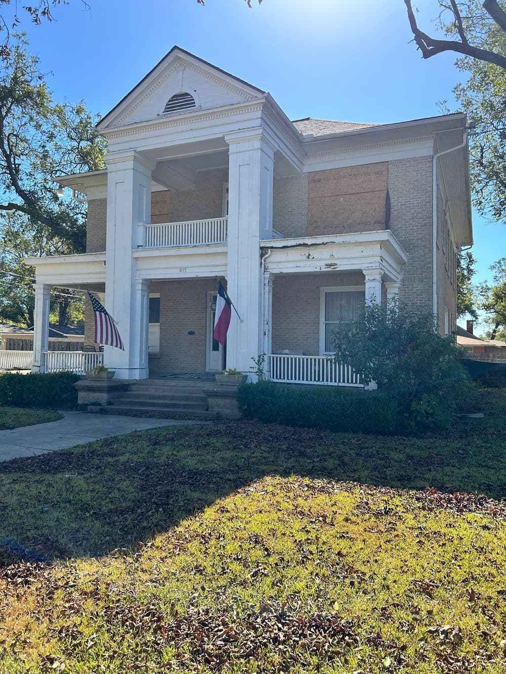 Historic two-story home with Texas and American flags, surrounded by lush greenery.
