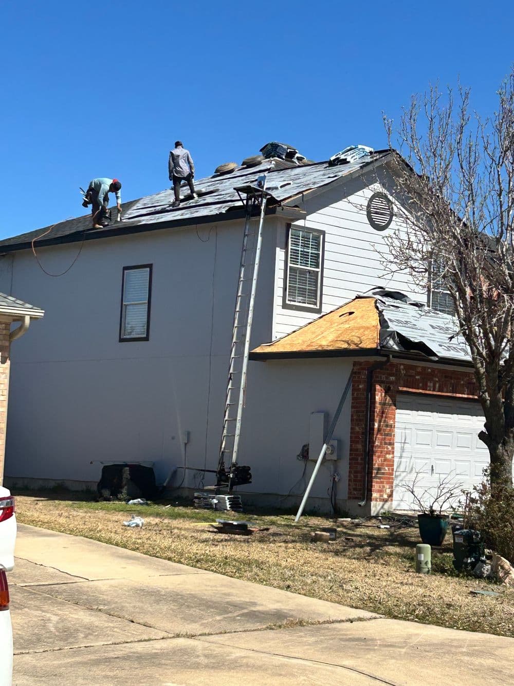 Roofers working on a residential home roof repair with ladder and tools under clear blue sky.