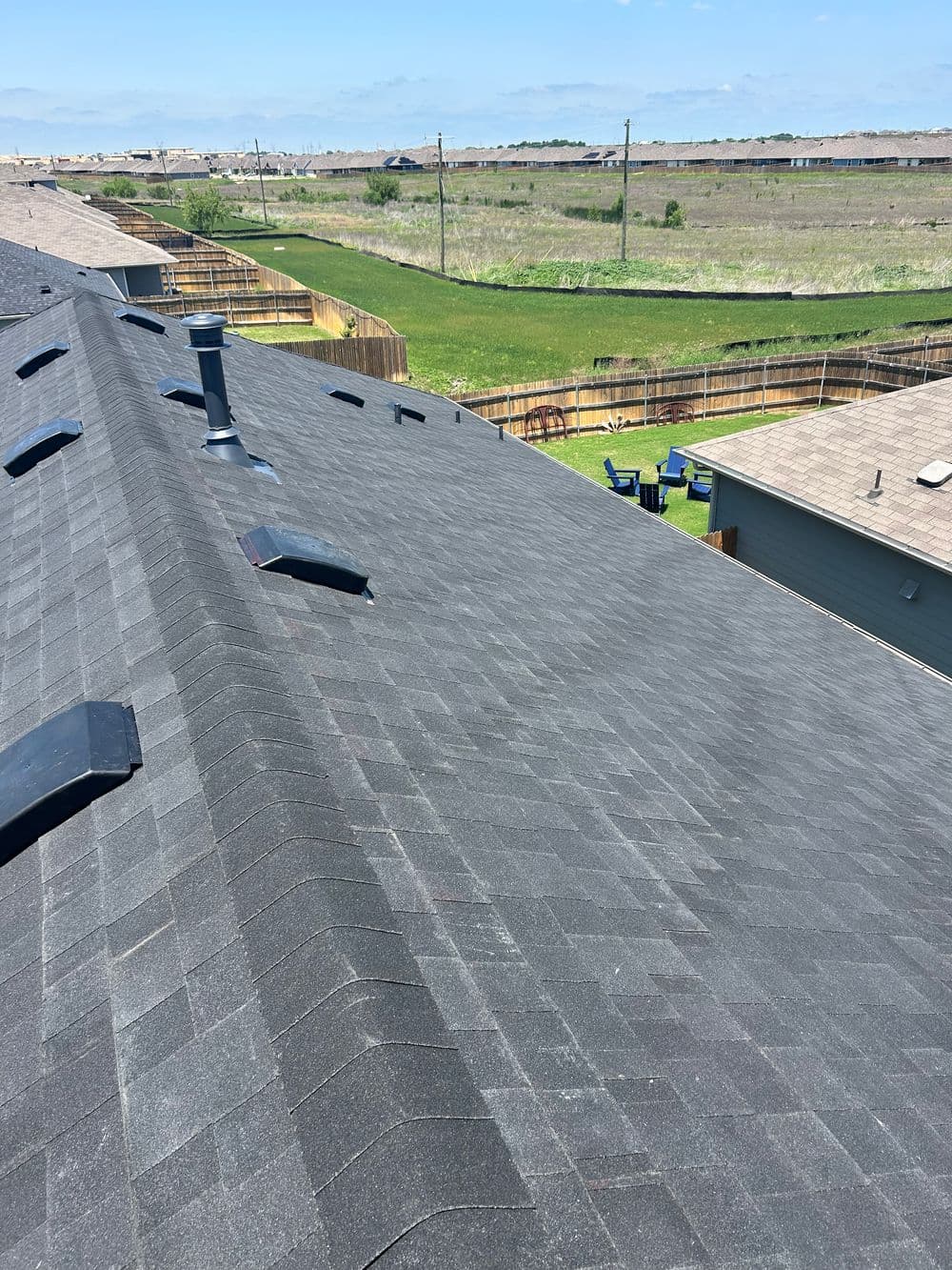 Roof view with grey shingles, skylights, and green backyard landscape under a clear blue sky.