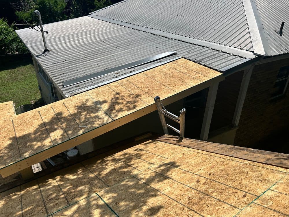Roofing work in progress with plywood sheets laid on a sloped roof. Ladder leaning against the building.