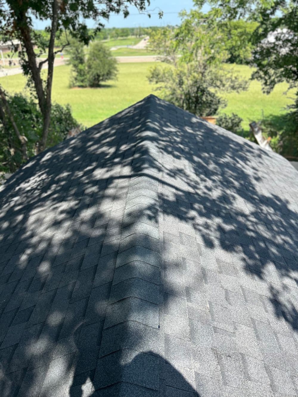 Aerial view of a gray shingled roof with trees and a green landscape in the background.