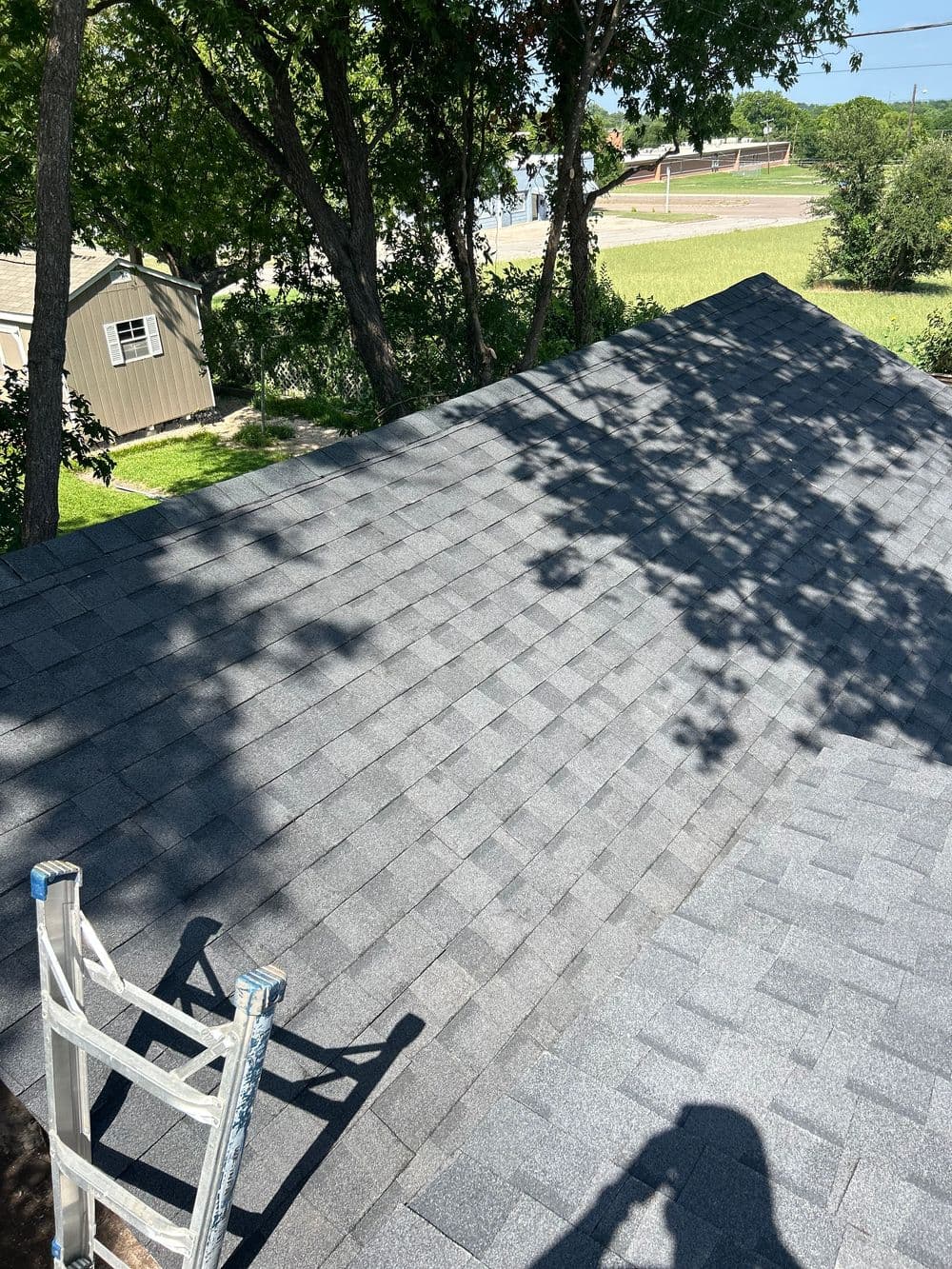 Gray shingle roof viewed from above, surrounded by trees and a clear blue sky.