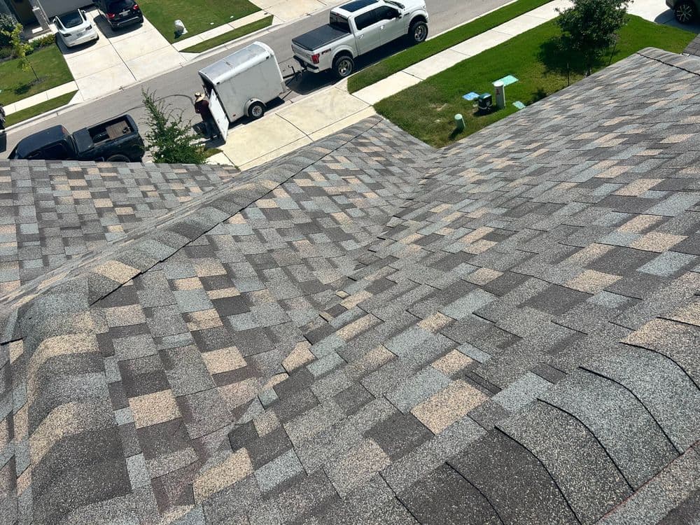 Aerial view of a shingled roof with uneven patches and nearby vehicles in a residential area.