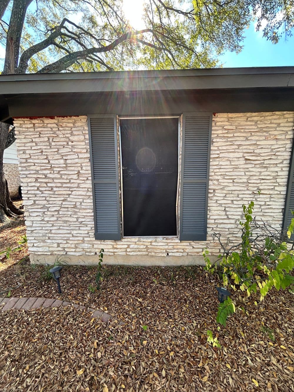 Black window covering with shutters on a textured stone house exterior, surrounded by leaves.