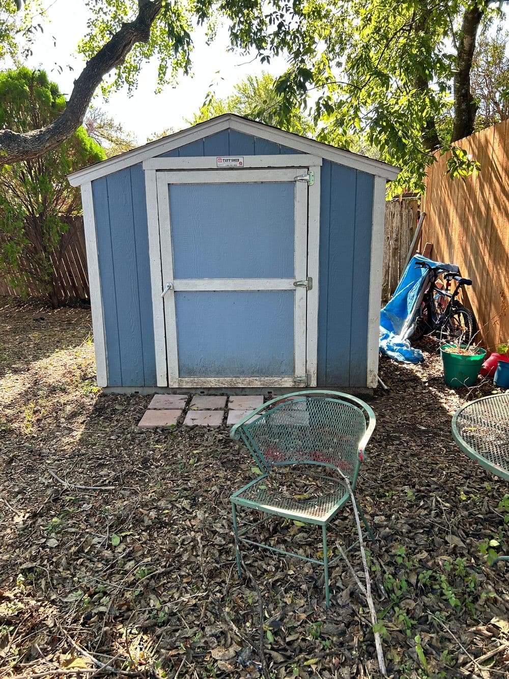Blue backyard shed with a white door, surrounded by chairs and plants.