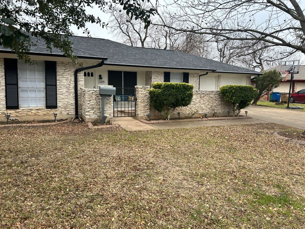 Single-story stone house with a lawn, mailbox, and trees on a cloudy day.