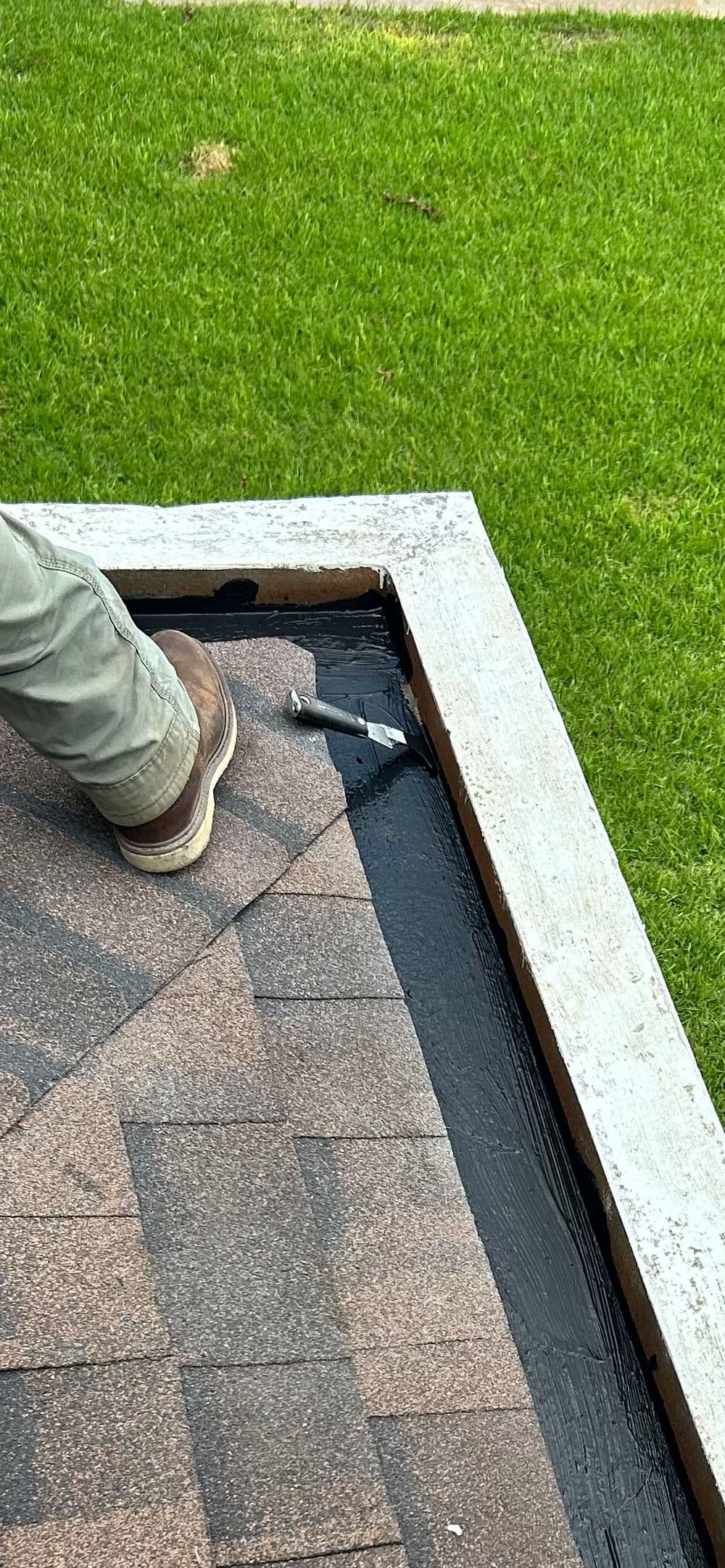 Person applying sealant on a roof edge with brown shingles and green grass in the background.