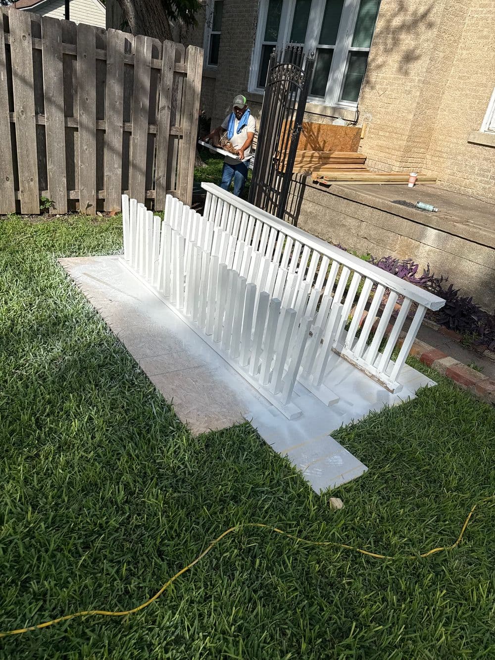 White picket fence being painted in a garden, with a person working in the background.