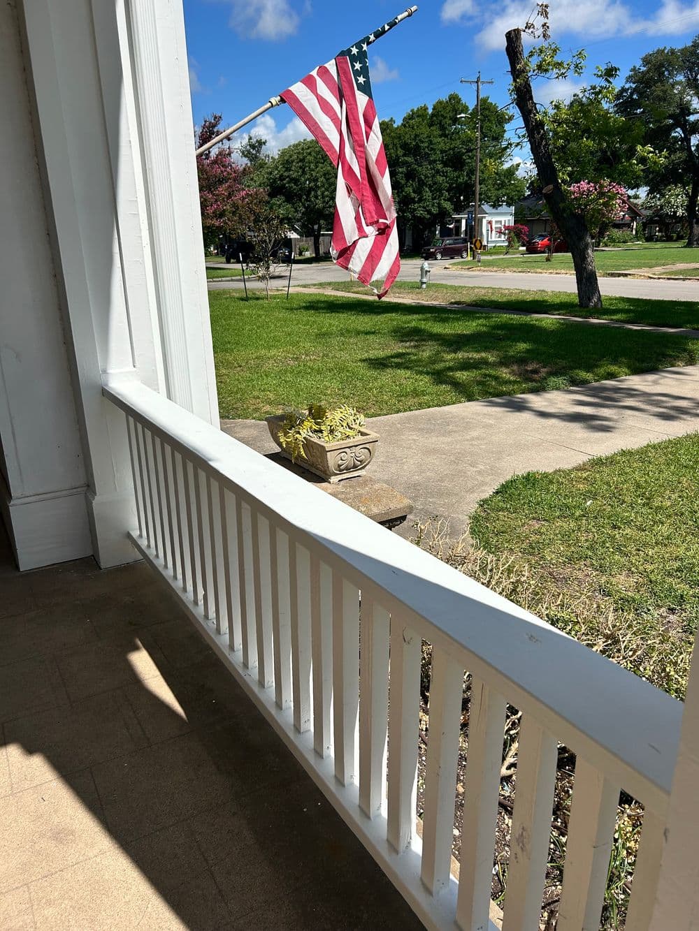 American flag waving in the breeze, view from a white porch overlooking a green yard.
