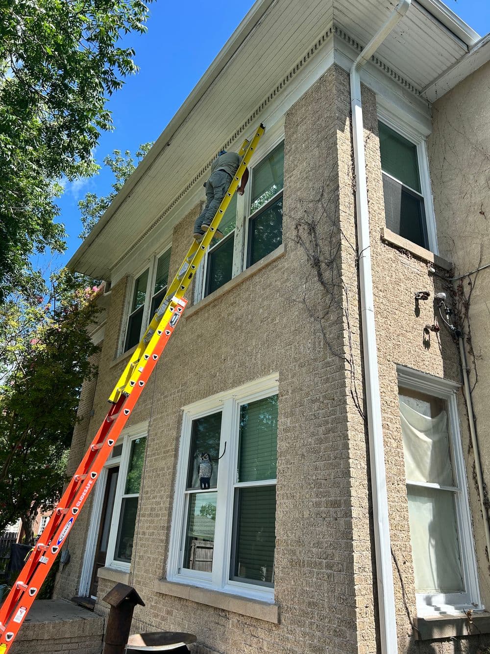 Person using a ladder to paint the exterior of a brick building on a sunny day.