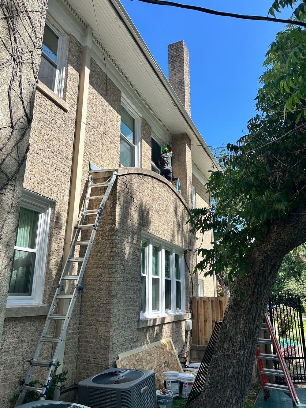 Worker using a ladder to paint the side of a house under a clear blue sky.