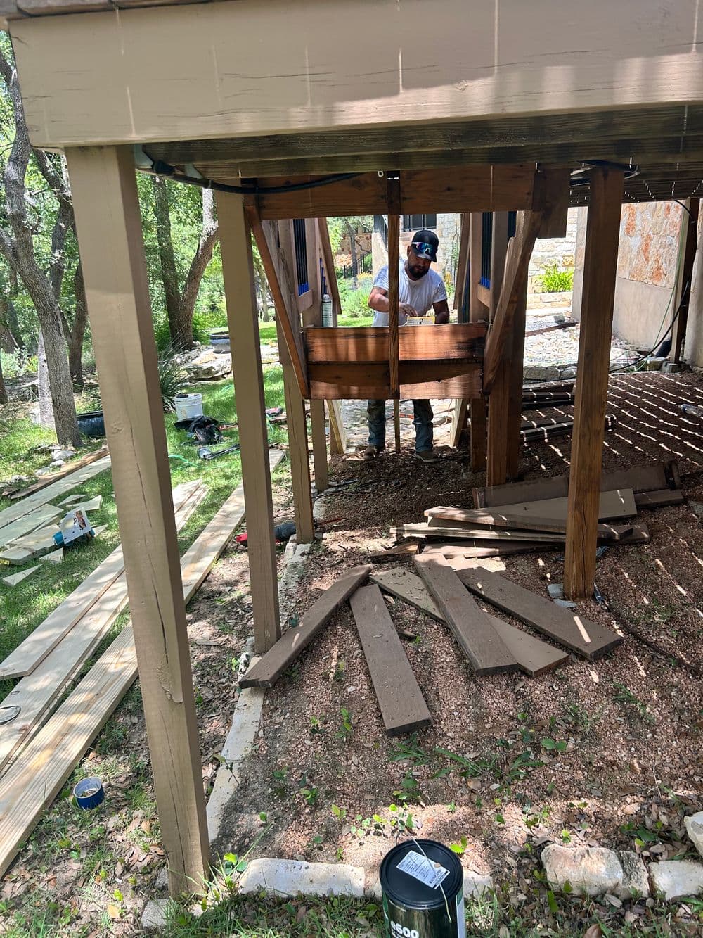 Construction worker repairing a wooden deck under a house surrounded by greenery.