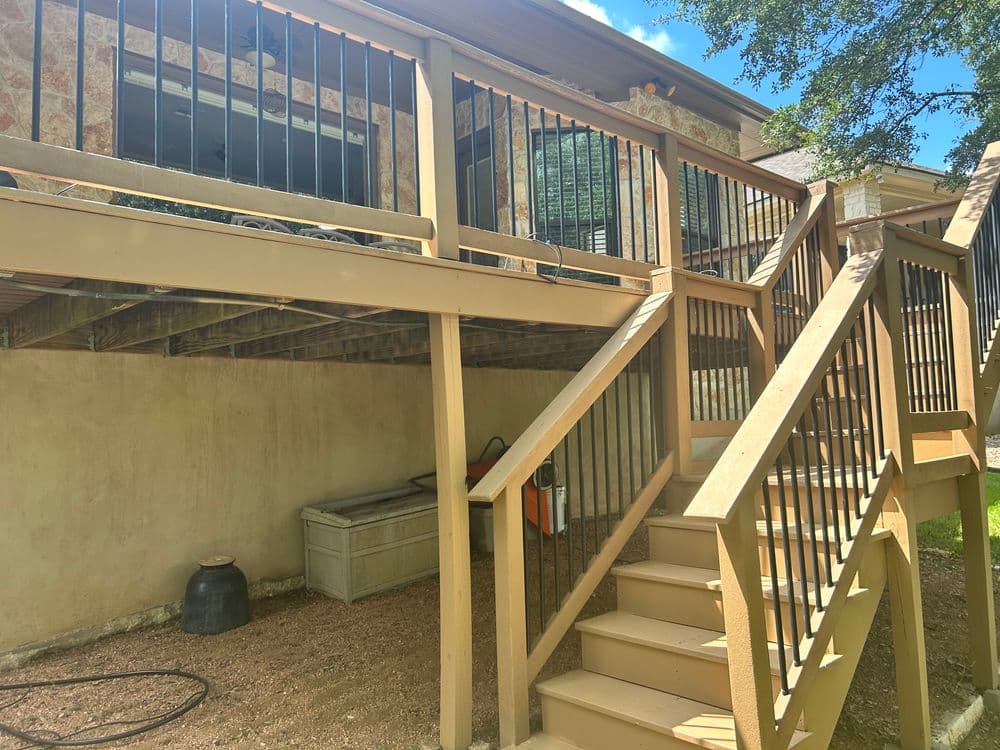 Wooden deck with black railing and staircase, leading to a house with a garden area below.