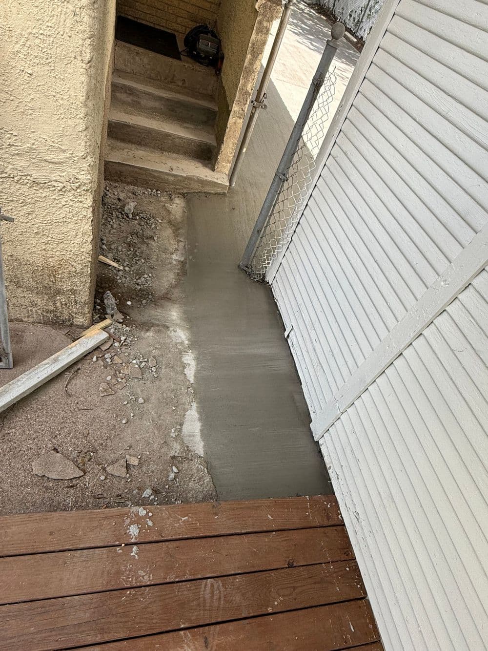 Newly poured concrete pathway beside a wooden deck and gate, showing construction progress.