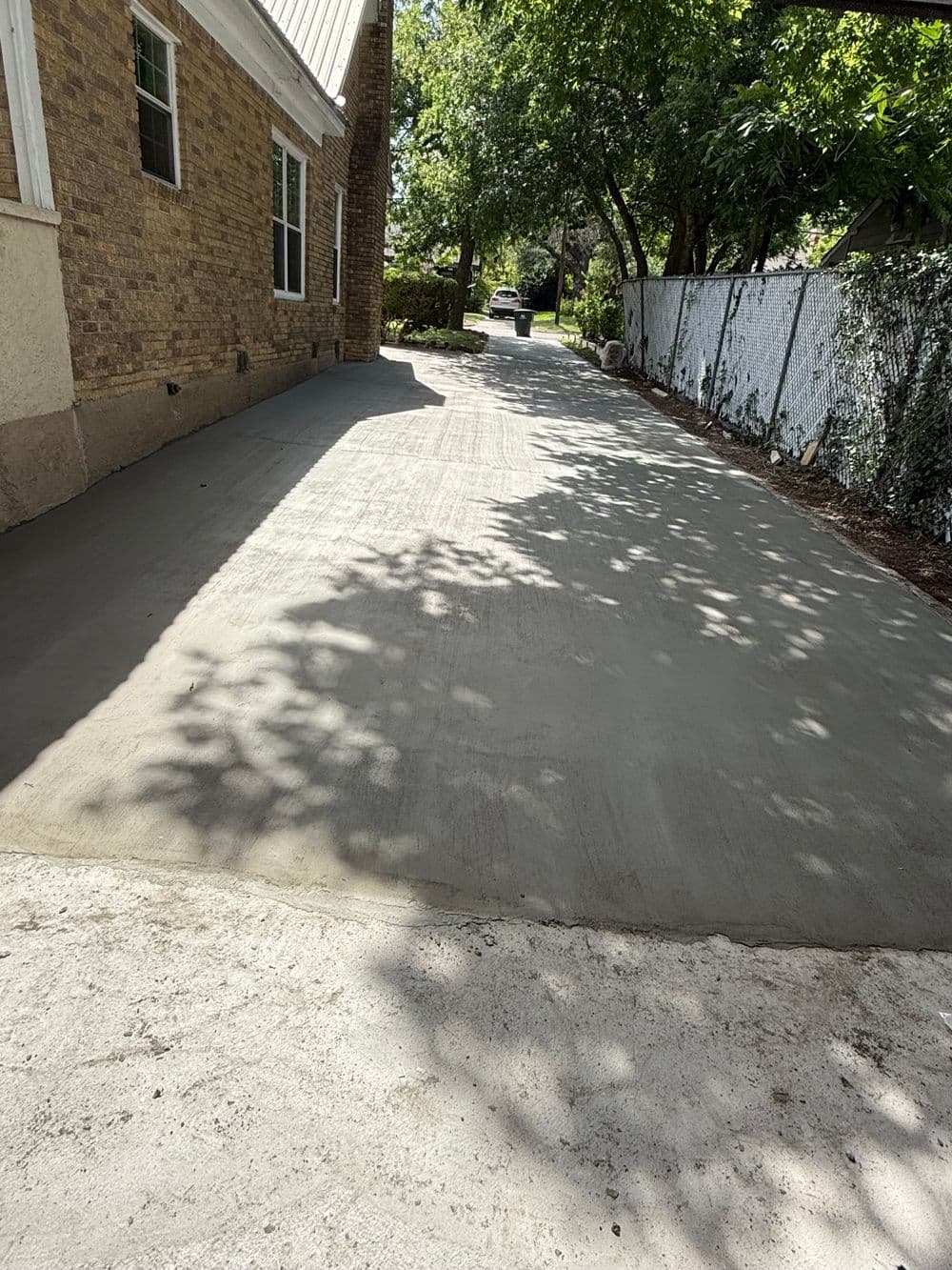 Freshly poured concrete driveway beside a house, surrounded by greenery and a fence.